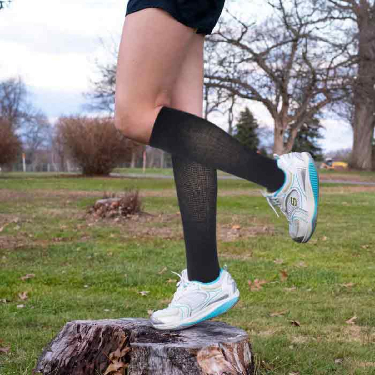 Person wearing black Dr. Motion knee-high socks and white sneakers on a grassy area with trees in the background